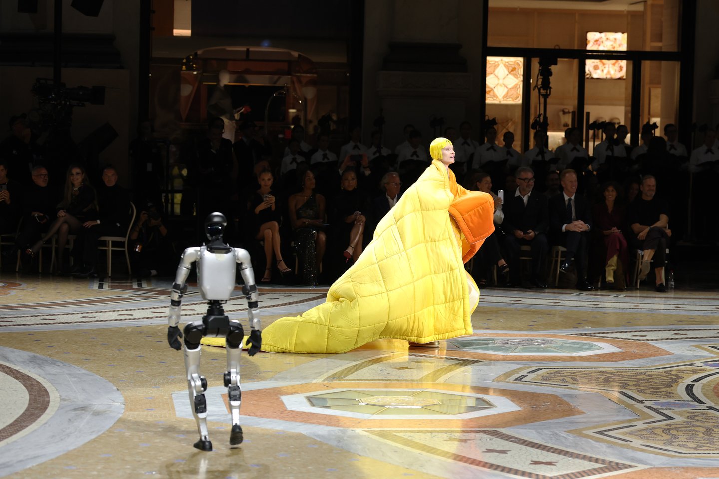 A humanoid robot and a model in a sculptural yellow look at Vogue World Milano 2026.