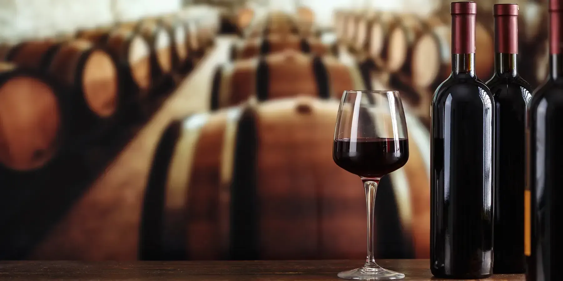 Ein Glas Rotwein neben dunklen Flaschen, dahinter Eichenfässer im Weinkeller.
