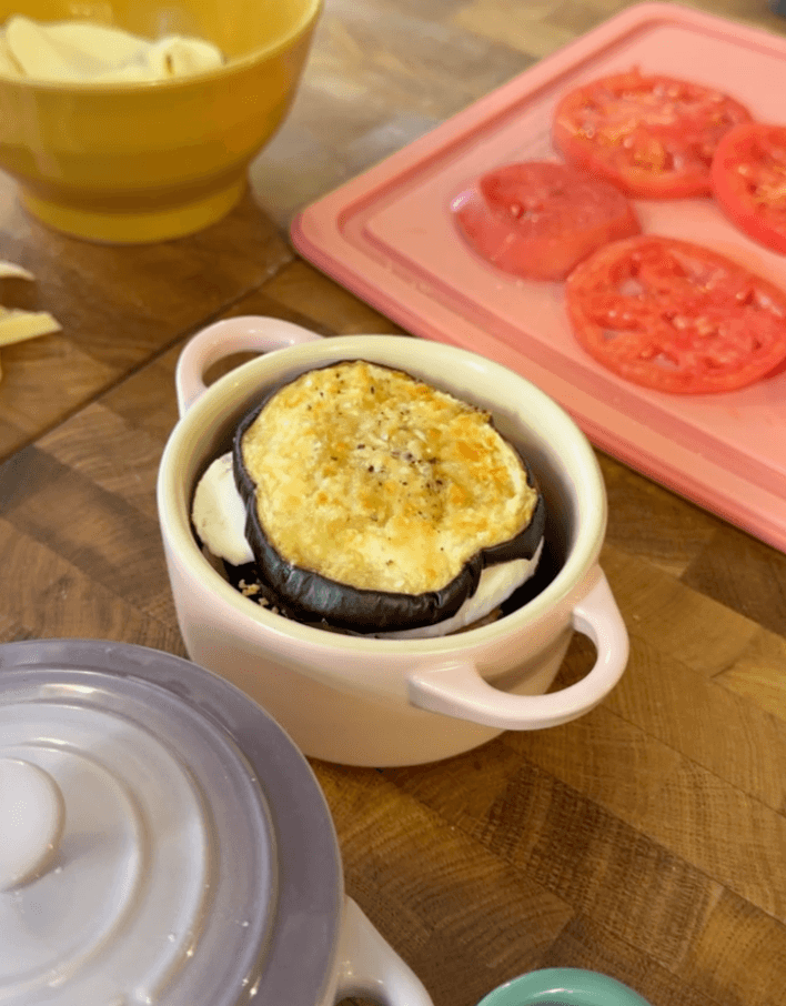 Parmigiana in una piccola cocotte rosa chiaro, con fette di pomodoro accanto.