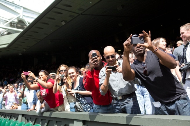 Espectadores levantan teléfonos y cámaras junto a una pista de tenis en Wimbledon.